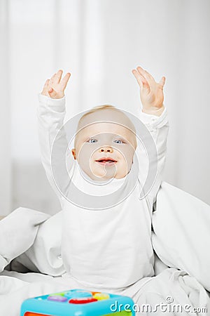 Frisky baby in crawlers sits on bed with raised hands Stock Photo