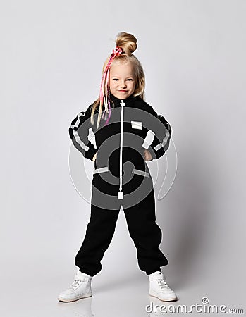 Frolic blonde kid baby girl with braids in black jumpsuit and sneakers stands with hands on waist and makes grimaces Stock Photo