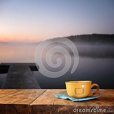 Front image of coffee cup over wooden table in front of calm foggy lake view at sunset Stock Photo