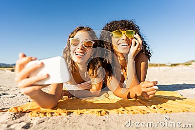 Front portrait of two cute girls with sunglasses in summer vacation using smartphone taking a selfie lying on sea sand of a Stock Photo