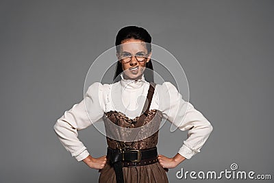 View of angry steampunk woman in glasses standing with hands on hips on grey Stock Photo