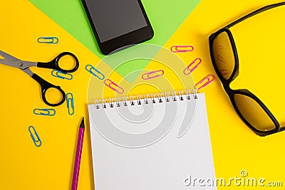 Front view colored blank paper sheet clips pencil smartphone scissors eyeglasses spiral notepad color background. Empty Stock Photo