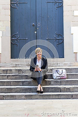 Front view of a elegant smiling business woman sitting on stairs outdoors while looking away holding a take away coffee in a sunny Stock Photo