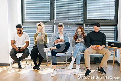 Front view of group five young diverse ethnicity candidates using mobile phones while waiting job interview in modern Stock Photo