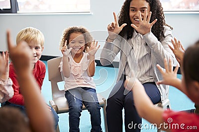 Front view of infant school children sitting on chairs in a circle in the classroom, holding up their hands and learning to count Stock Photo