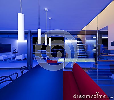 Front view living room with table and a red sofa lit by blue LEDs and lamps attached to the ceiling. Large window overlooking the Stock Photo