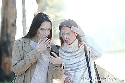 Two worried women checking phone news in a park Stock Photo
