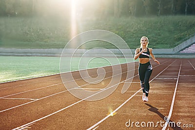 Front view of running caucasian woman in black sportswear doing cardio workout on sports track of stadium. Adult female Stock Photo