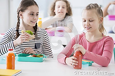 Front view of two junior girls in a school cafeteria during lunch break. One holding a wholewheat sandwich, the other opening a b Stock Photo