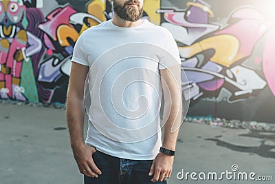 Front view. Young bearded hipster man dressed in white t-shirt is stands against wall with graffiti. Mock up. Stock Photo