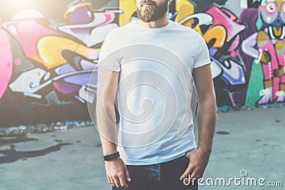 Front view. Young bearded hipster man dressed in white t-shirt is stands against wall with graffiti. Mock up. Editorial Stock Photo