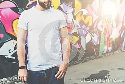 Front view. Young bearded hipster man dressed in white t-shirt is stands against wall with graffiti. Mock up. Editorial Stock Photo