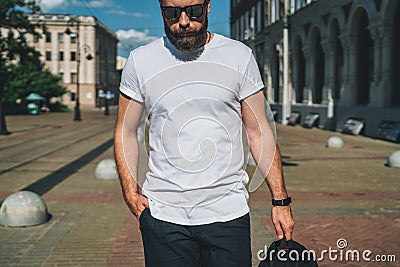 Front view. Young bearded millennial man dressed in white t-shirt and sunglasses is stands on city street. Mock up. Stock Photo