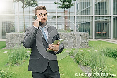 Young smiling businessman in suit and tie is standing outdoor,holding tablet computer and talking on his cell phone Stock Photo