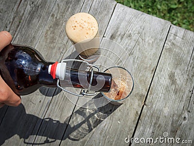 Frothy dark beer pouring into tall glasses from a brown glass bottle in summer garden on rustic wooden table Stock Photo