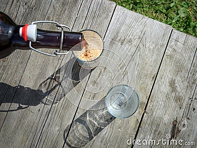 Frothy dark beer pouring into tall glasses from a brown glass bottle in summer garden on rustic wooden table Stock Photo