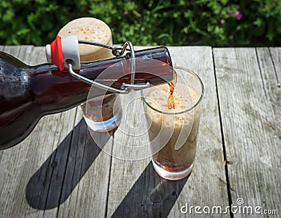 Frothy dark beer pouring into tall glasses from a brown glass bottle in summer garden on rustic wooden table Stock Photo