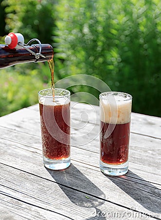 Frothy dark beer pouring into tall glasses from a brown glass bottle in summer garden on rustic wooden table Stock Photo