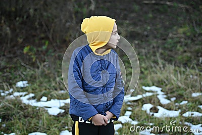 Frozen little thin caucasian girl in yellow hoodie and blue jacket shiver near snow on ground in outdoor park during leisure Stock Photo