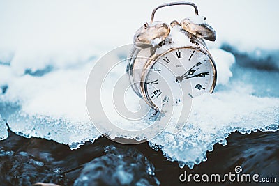 Frozen time. A clock on an ice next to the water. Extreme weather situation. Snow falling on a clock in a nature. Stock Photo