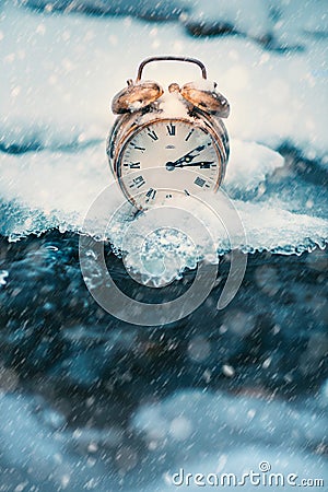 Frozen time. A clock on an ice next to the water. Extreme weather situation. Snow falling on a clock in a nature. Stock Photo