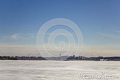 Frozen white snow lake on the background of the city with high-rise buildings and smoking huge pipes under a clear blue sky Stock Photo