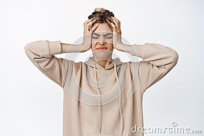 Frustrated blond girl holding hands on head and close eyes, grimacing upset, standing troubled against white background Stock Photo