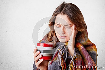 Frustrated ill young woman covered in warm blanket, keeps hand on temple, suffers from terrible headache, drinks hot beverage, tri Stock Photo