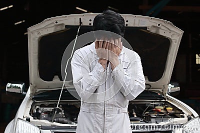 Frustrated stressed young mechanic man in white uniform covering his face with hands against car in open hood at the repair garage Stock Photo