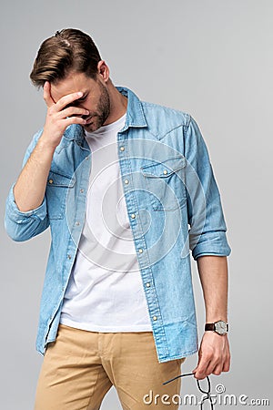 Frustrated young caucasian man in jeans shirt touching head with hand while standing overg studio grey background Stock Photo