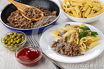 Frying pan with fried mince, bowl with pasta, green peas, ketchup, plate with macaroni, fried mincemeat, fork on table Stock Photo