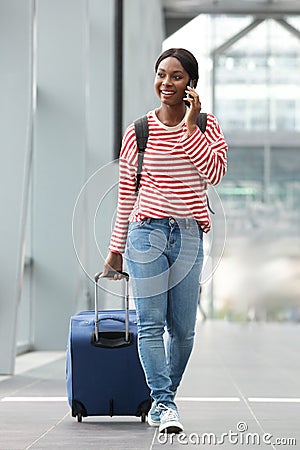 Full body happy young black woman walking with suitcase in airport terminal with cellphone Stock Photo