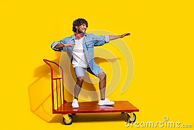 Full body photo of attractive man ride warehouse cart wink dressed stylish denim clothes red scarf isolated on yellow Stock Photo