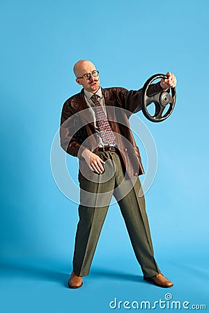 Full body shoot of young balded and mustache man holding car wheel over blue studio background. Vertical photo. Stock Photo