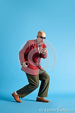 Full lenght image. Balded man with mustache dressed in business retro suit sneaking over blue studio background. Stock Photo