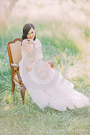 The full-lenght photo of the smiling woman in long white dress sitting on the old modern chair in the green spring field Stock Photo