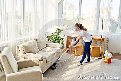 Full length body portrait of young woman in white shirt and jeans cleaning carpet with vacuum cleaner in living room, copy space. Stock Photo