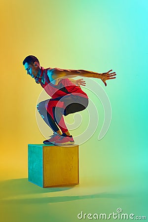 Full-length dynamic image of y9ung muscular man training, jumping on block, training against gradient blue yellow Stock Photo
