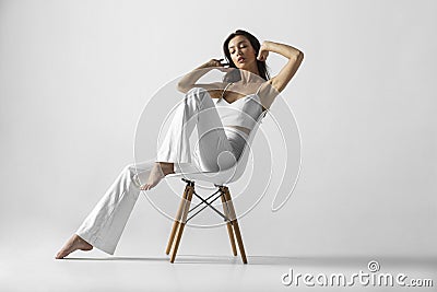 The full length image of a young brunette woman with makeup and hairstyle, dressed in white, sits relaxed on the chair. Stock Photo