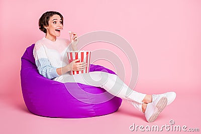 Full length photo portrait of excited girl eating popcorn sitting in violet beanbag chair isolated on pastel pink Stock Photo