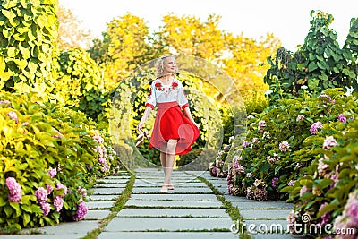 Full length portrait of happy barefoot attractive woman in stylish red white dress holding shoes and walking on tile path in Stock Photo