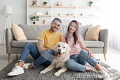 Full length portrait of positive international couple with their pet dog sitting on soft carpet at home Stock Photo