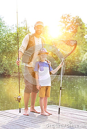 Full length portrait of smiling father and son standing with fishing tackles on pier against lake Stock Photo