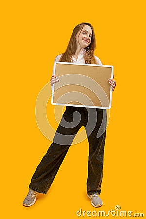 Full-length portrait of smiling young woman with note board or cork board, yellow background. Copy space Stock Photo