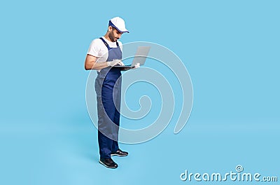 Full length professional workman in uniform and protective gloves typing on laptop keyboard, online communication Stock Photo