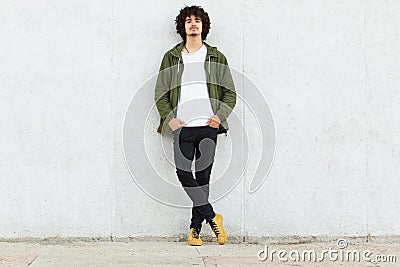 Full length shot of serious unshaven guy wears white t shirt, green anorak, black trousers and sneakes, keeps hands in pockets, ha Stock Photo