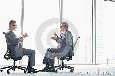 Full-length side view of businessmen discussing while sitting on office chairs by window Stock Photo