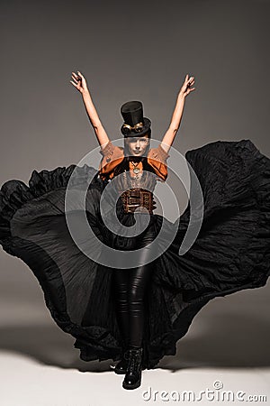 Length view of attractive steampunk woman in top hat with goggles standing with hands up on grey Stock Photo