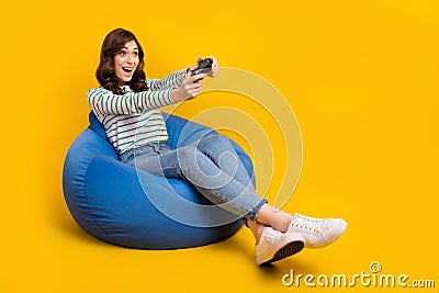 Full size photo of cheerful woman striped sit on armchair look empty space hold joystick play game isolated on yellow Stock Photo