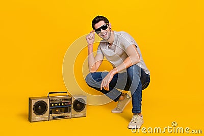 Full size photo of nice brunet young guy sit near boombox wear t-shirt glasses jeans sneakers on yellow Stock Photo